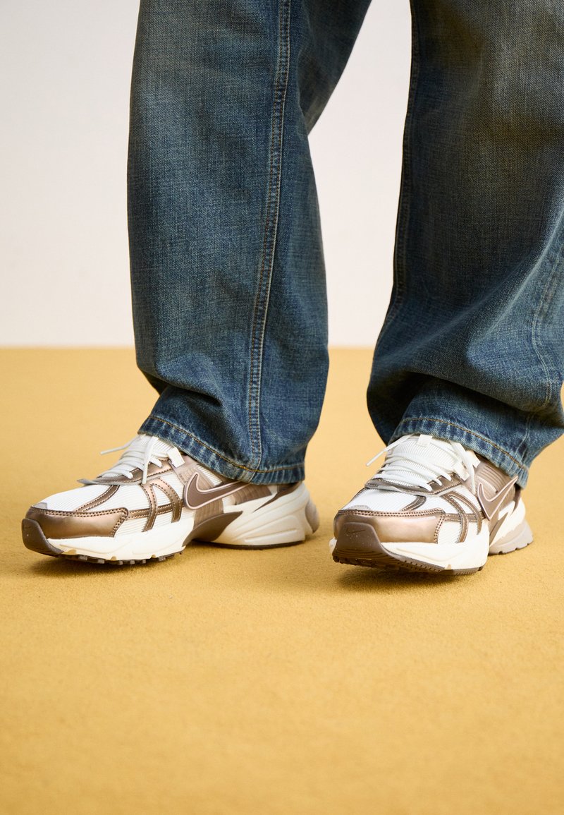 Zapatillas de running Nike en blanco y bronce metálico, que cuentan con materiales de malla y sintéticos, con una suela texturizada y un diseño de cordones.