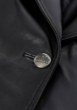 Zbliżenie na czarną skórzaną kurtkę z srebrnym guzikiem, na którym wygrawerowano dwa lwy, koronę oraz nazwę marki "WITTCHEN".
