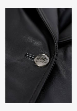 Zbliżenie na czarną skórzaną kurtkę z srebrnym guzikiem, na którym wygrawerowano dwa lwy, koronę oraz nazwę marki "WITTCHEN".