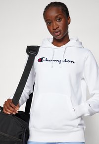Hvit bomullshoodie med lommer foran, med marineblå brodert "Champion"-logo. Brukt med en svart veske-rem over skulderen.