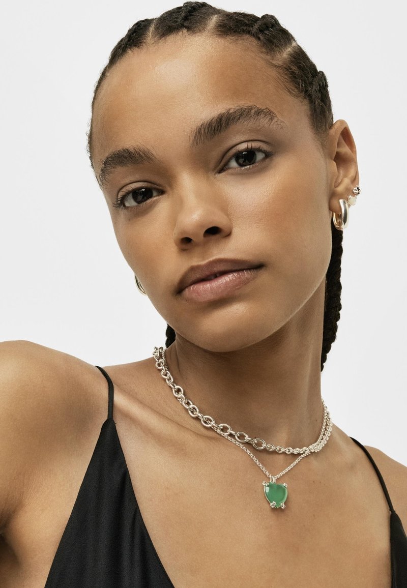 Collane in argento a strati, con un pendente verde. Top nero a spaghetto, sfondo minimale, che mette in risalto i dettagli e il design dei gioielli.