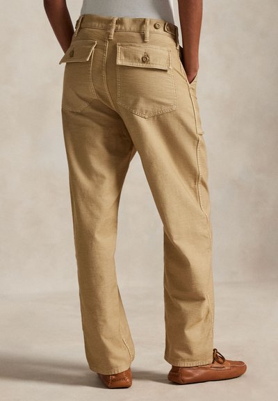 Person wearing beige straight-leg pants with buttoned back pockets and brown moccasin-style shoes, standing with hands in side pockets.