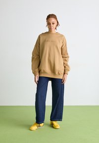 Rudas džemperis su iškilmingu logotipu, per didelis pasiuvimas ir pūkuotos rankogaliai. Suderintas su plačiais tamsiais džinsais ir geltonais sportbačiais su juodomis juostomis.