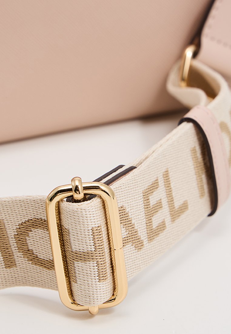 Sangle en tissu beige réglable avec un détail de boucle en or, attachée à un sac en cuir rose clair en arrière-plan.