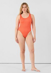 Stressige oranje ribbed badpak met brede bandjes en hoge beenuitsnijdingen. Het model draagt witte slippers tegen een effen achtergrond.