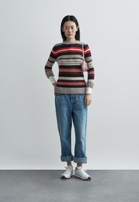 Knit sweater in stripes of red, brown, and cream, paired with light blue jeans and white sneakers. Features a ribbed hem and cuffs.
