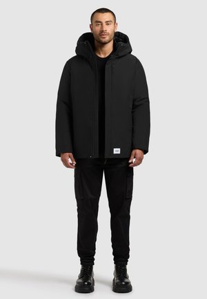 Schwarze Kapuzenjacke mit glatter Textur, Reißverschluss und Seitentaschen, kombiniert mit schwarzen Cargo-Hosen und robusten schwarzen Stiefeln.