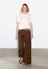 Light pink cotton t-shirt, loose-fitting brown linen pants with a belt, and black flip-flops, against a plain white background.