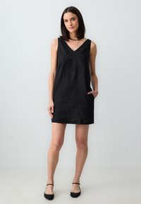 Schwarzes ärmelloses Kleid mit V-Ausschnitt, mit Taschen und lockerer Silhouette. Aus leichtem Material, glatter Textur, knielang.