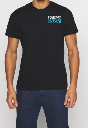Homme portant un t-shirt noir à manches courtes avec un logo blanc et bleu "TOMMY JEANS" sur le côté gauche de la poitrine, associé à un jean bleu foncé.