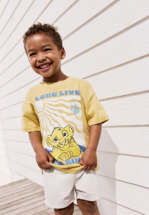 Žluté grafické tričko s motivem lva, textem "AŤ ŽIJE KRÁL", krátké rukávy, v kombinaci s lehkými šortkami, stojící před bílým dřevěným pozadím.