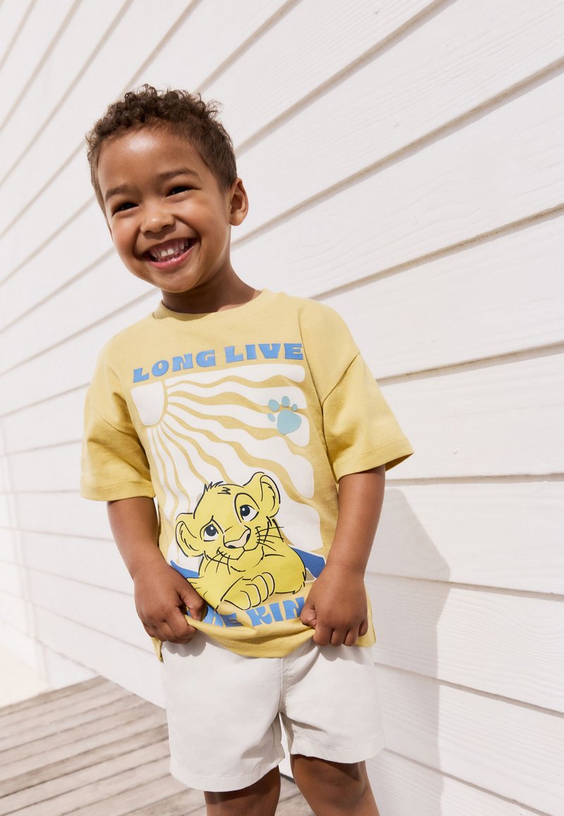 Sárga grafikai póló oroszlános mintával, „LONG LIVE THE KING” felirattal, rövid ujjú, világos rövidnadrággal párosítva, fehér fa háttér előtt állva.