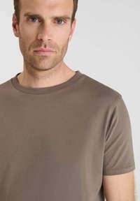 Uomo con capelli corti e marroni e barba, che indossa una maglietta girocollo marrone semplice su uno sfondo chiaro e uniforme.