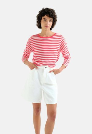 Persona joven con cabello rizado que lleva una camisa de manga larga a rayas rojas y blancas y pantalones cortos blancos de tiro alto, de pie con las manos en los bolsillos.