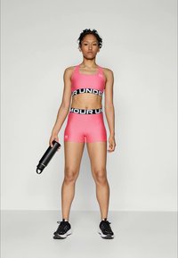 Γυναίκα με ροζ αθλητικό σουτιέν και σορτς κρατώντας ένα μαύρο μπουκάλι νερού, στέκεται μπροστά από ένα απλό φωτεινό φόντο φορώντας μαύρα αθλητικά παπούτσια.