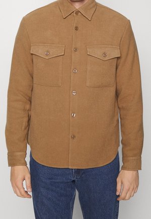 Man draagt een kamelkleurig wollen overhemdjack met knopen, twee borstflappen en een blauwe spijkerbroek tegen een neutrale achtergrond.