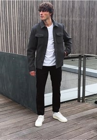 Giacca grigia con bottoni e due tasche sul petto sopra una t-shirt bianca, abbinata a pantaloni neri e sneaker bianche, in piedi su un ponte di legno.