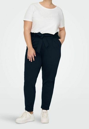 Person trägt ein weißes kurzärmeliges Hemd, schwarze hoch taillierte Hose mit gebundenem Gürtel und weiße Turnschuhe und steht mit einer Hand in der Tasche.