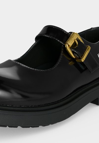 Chaussure Mary Jane en cuir verni noir avec semelle épaisse et boucle dorée gravée du nom de la marque.