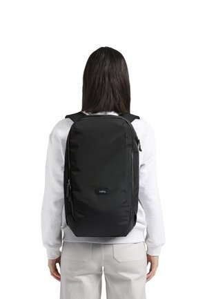 Sac à dos noir avec une finition lisse et texturée. Présente un design épuré, des compartiments zippés et une étiquette de logo. Porté sur les épaules.