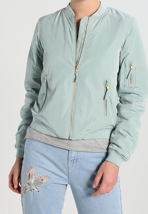 Personne portant une veste bomber vert clair avec des fermetures éclair dorées et un jean bleu clair avec un oiseau brodé sur la cuisse gauche.
