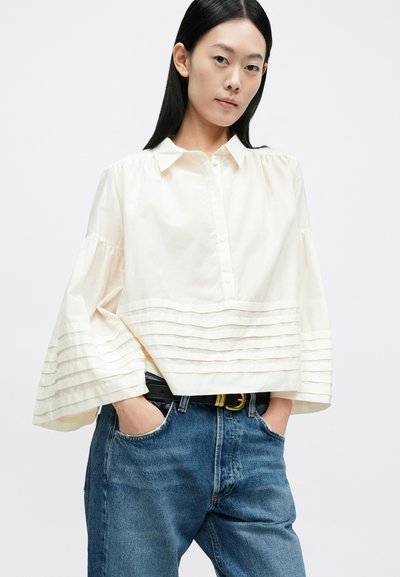 Kremfarvet skjorte med knapper og plisseret design ved kant og ærmer, kombineret med højtaljede blå jeans og sort bælte. Ensfarvet baggrund.