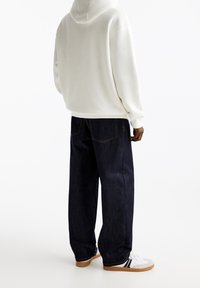 Persoon in een witte hoodie, donkere, loszittende jeans en witte sneakers met zwarte strepen, staand tegen een plain witte achtergrond.