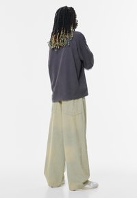 Felpa oversize grigia scura abbinata a pantaloni in denim leggeri di un giallo-verde tenue. Tessuto morbido, vestibilità rilassata e dettagli visibili delle tasche posteriori.