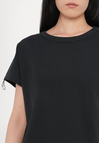 Svart T-skjorte med korte ermer og rund hals. Laget av et glatt stoff, med en subtil glans og dekorativ kant langs ermene.