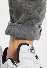 Balti Nike Air Force 1 sportbačiai su juodomis akcentais, derinami su pilkos džinsinės kelnės, turinčiomis sukurtą apvyniotą kraštą ir oranžinę siuvinėjimo detalę.