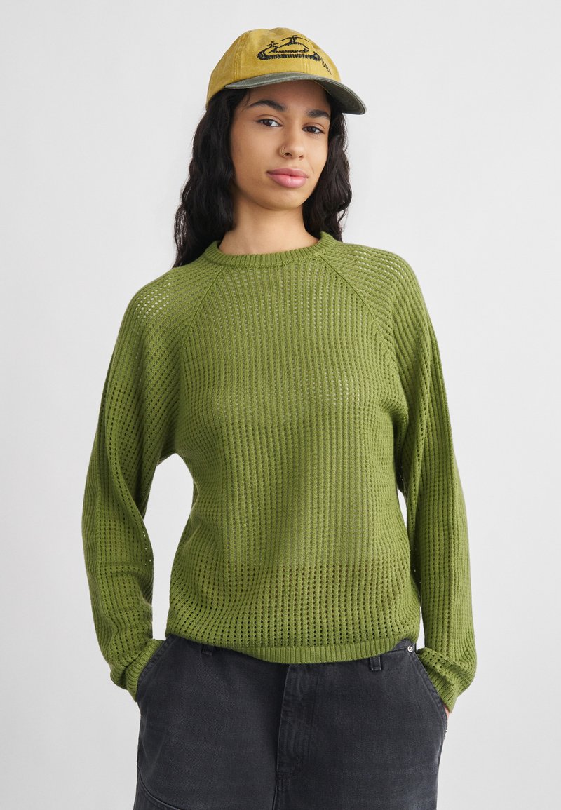 Γυναίκα με μακριά σκουρόχρωμα σγουρά μαλλιά, που φορά κίτρινο καπέλο, πράσινο πλεκτό πουλόβερ και σκούρα παντελόνια, με τα χέρια στις τσέπες, στέκεται μπροστά σε λευκό φόντο.