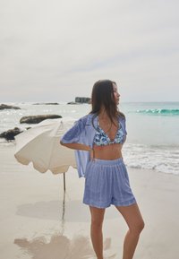 Světlé modré texturované šortky a ladící otevřená košile přes potisknuté bikiny, stojící na pláži s béžovým slunečníkem v pozadí.