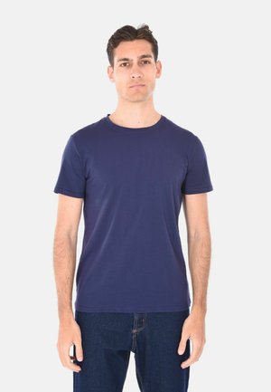 Giovane uomo in piedi contro uno sfondo uniforme, indossa una t-shirt blu navy a maniche corte e jeans blu scuro, guarda avanti con un'espressione neutra.