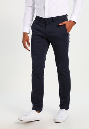 Mann trägt marineblaue Slim-Fit-Hose, weißes Knopfbügeleisenhemd und weiße Turnschuhe, steht mit einer Hand in der Tasche.
