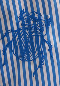 Il tessuto a righe blu e bianche presenta un ricamo blu dettagliato con un design di scarabeo, evidenziando un lavoro di linea intricato e texture.