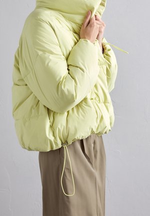 Personne portant une doudoune vert clair avec cordons, tenant le col près, associée à un pantalon marron ample sur fond uni.