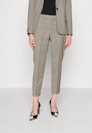 Person trägt hellgraue, maßgeschneiderte, kurze Hosen und Blazer mit schwarzem Top und schwarzen spitzen High Heels mit metallischen Spitzen.