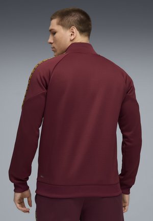 Bordeaux atletisk jakke med høj krave, lange ærmer og gyldne detaljer langs skuldrene. Glat tekstur og tætsiddende design.