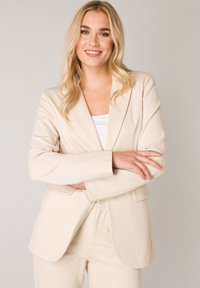 Beige op maat gemaakte blazer met klepels, sluiting met één knop, twee voorzakken en een zachte textuur, gecombineerd met bijpassende broek.