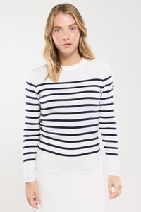 Vrouw met blond haar, gekleed in een witte trui met horizontale marineblauwe strepen en een witte rok, staand tegen een effen lichte achtergrond.