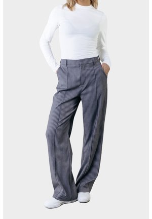 Woman wearing white long-sleeve top, gray pinstripe wide-leg trousers, white sneakers, standing with hands in pockets against plain background.