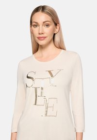 Jasnobeżowa koszula z długim rękawem z błyszczącym złotym nadrukiem "STYLE" z akcentami w formie kropek. Wykonana z miękkiej, gładkiej tkaniny.