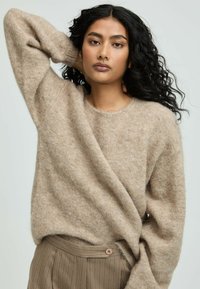 Frau mit langen, lockigen schwarzen Haaren, die einen beigen, flauschigen Pullover und braune gestreifte Hosen trägt, und mit einer Hand hinter ihrem Kopf posiert.