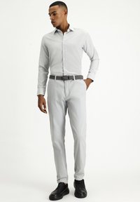 Camicia button-up grigia chiara a fantasia con maniche lunghe, abbinata a pantaloni grigi chiari e scarpe slip-on nere. Presenta una cintura intrecciata grigia.