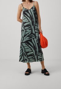 Vestido maxi sin mangas de estampado de hojas verdes con escote en V, combinado con un bolso texturizado naranja y sandalias de plataforma negras.