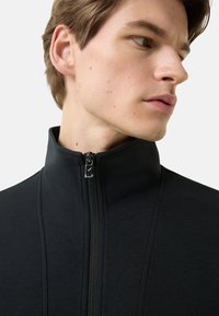 Czarna kurtka na zamek z wysokim kołnierzem i gładką fakturą z subtelnymi przeszyciami. Suwak posiada srebrny uchwyt.