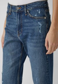 Jasnoniebieskie jeansy z denimu z wysokim stanem, z przetarciami, widocznymi szwami i pięcioma kieszeniami. Materiał ma teksturowane wykończenie.