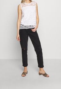 Vit ärmlös topp med spetsdetaljer och geometriska mönster, kombinerad med svarta rivna jeans och sandaler med leopardtryck.