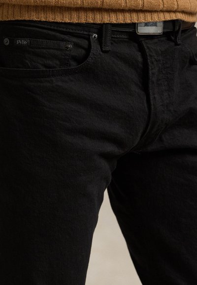 Fekete Polo farmer barna bordázott pulóverrel viselve, betűrve, amely egy csíkos mintájú övcsatot mutat.