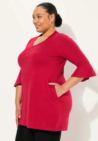 Tunic rossa realizzata in morbido tessuto, con scollo a V, maniche a tre quarti a campana e tasche laterali. Tessuto liscio e vestibilità ampia.
